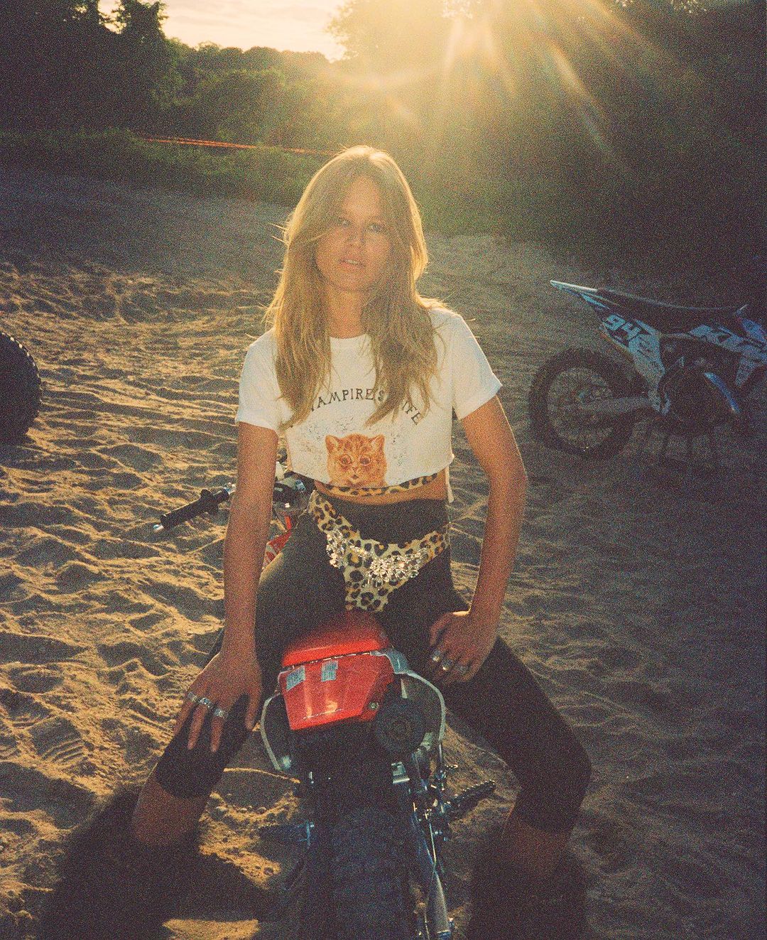 Blonde woman sitting on red dirt bike in sunset. The sun shines behind her in a sand dune area next to a green forest.