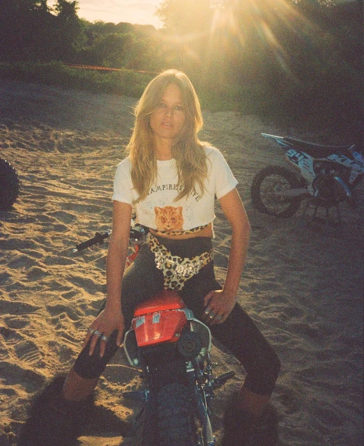 Blonde woman sitting on red dirt bike in sunset. The sun shines behind her in a sand dune area next to a green forest.