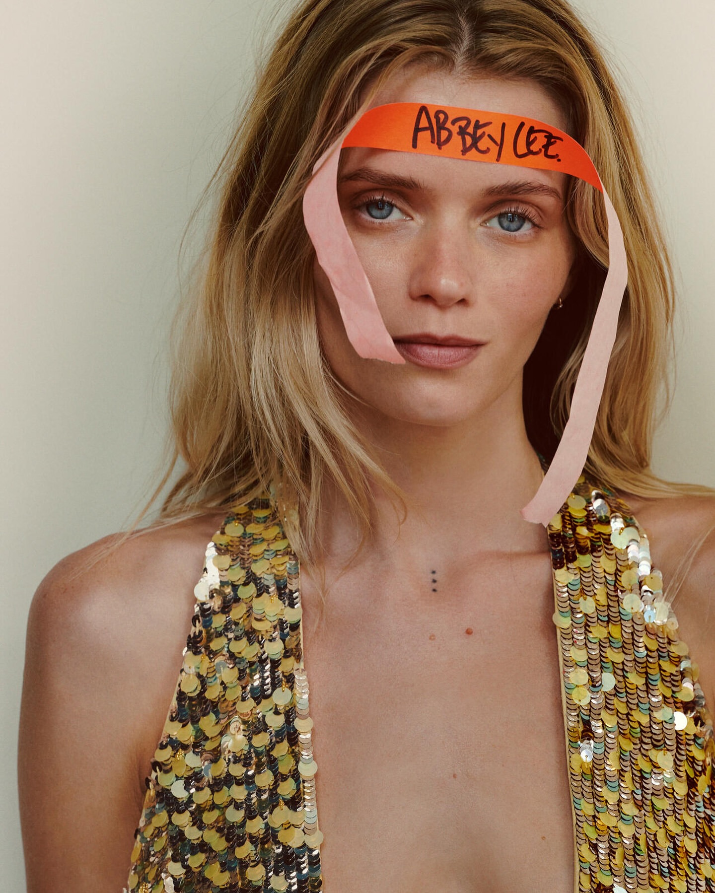 Close-up portrait of a blonde woman with an orange paper headband with handwritten text, wearing a sequined top and natural makeup.
