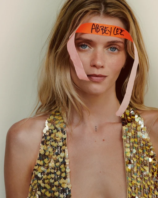 Close-up portrait of a blonde woman with an orange paper headband with handwritten text, wearing a sequined top and natural makeup.