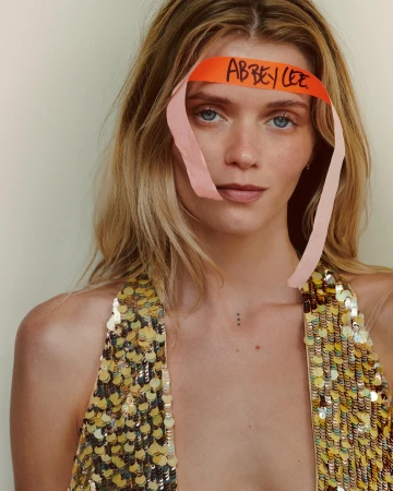 Close-up portrait of a blonde woman with an orange paper headband with handwritten text, wearing a sequined top and natural makeup.