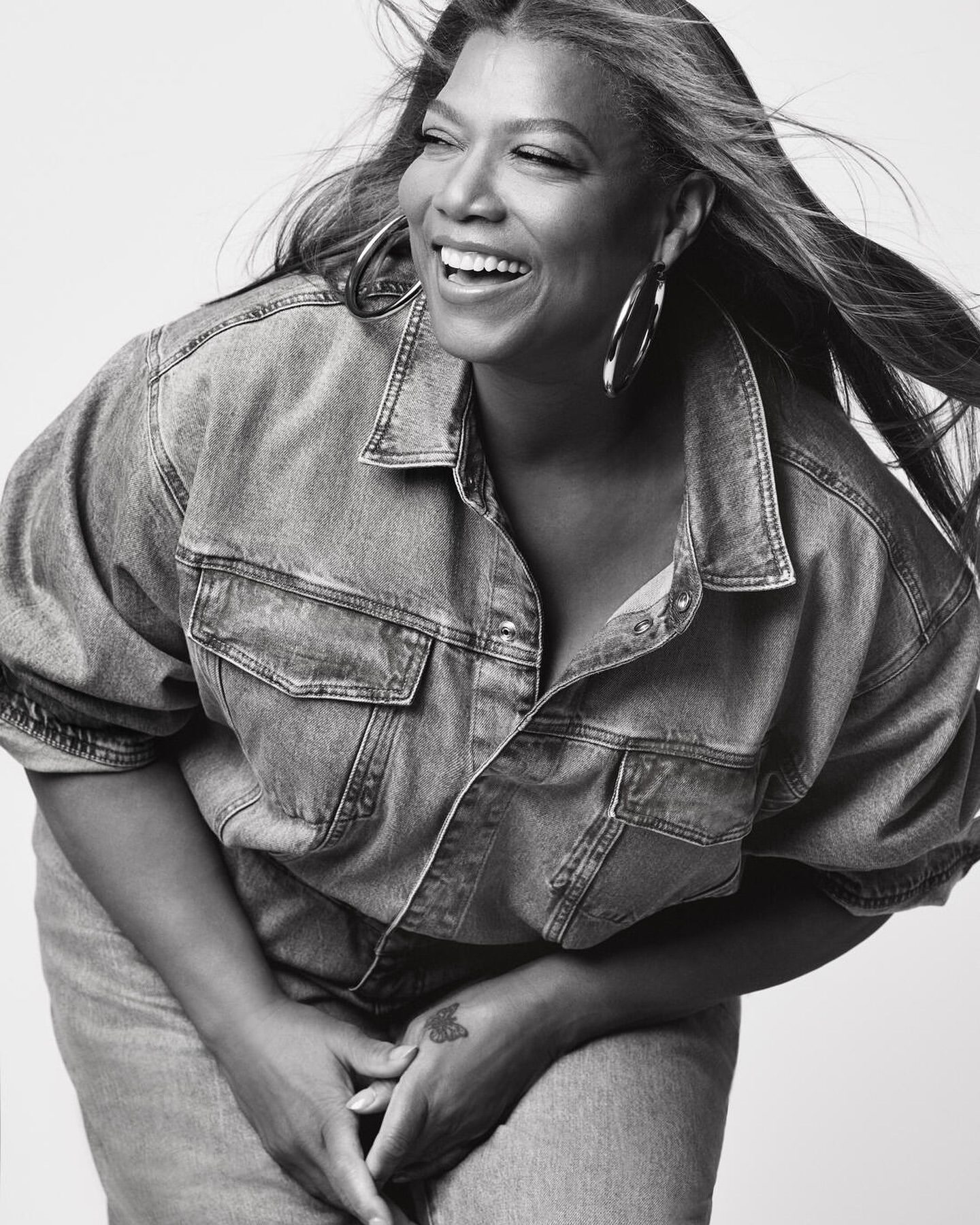Black and white portrait of smiling woman with denim jacket and hoop earrings, with windswept long hair, against a white backdrop.