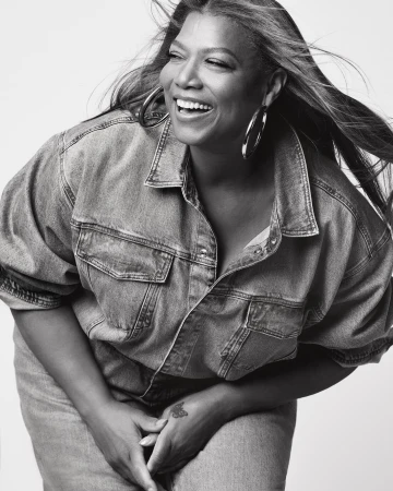 Black and white portrait of smiling woman with denim jacket and hoop earrings, with windswept long hair, against a white backdrop.