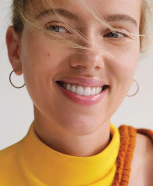 Close-up portrait of a smiling woman with blonde hair, wearing a yellow top and gold hoop earrings, set against a neutral background.