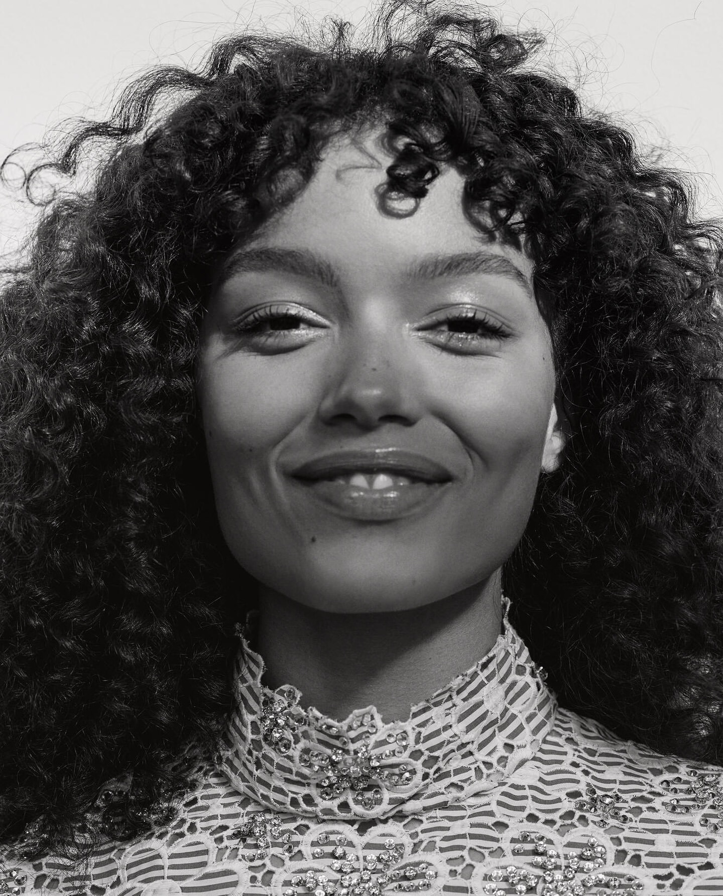 Close-up portrait of a smiling young woman with curly hair in a lace top, captured in a monochromatic, high-key lighting setup.