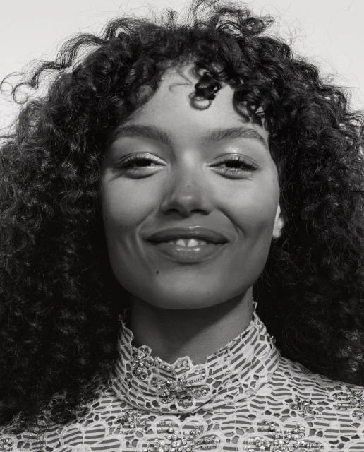 Close-up portrait of a smiling young woman with curly hair in a lace top, captured in a monochromatic, high-key lighting setup.