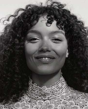 Close-up portrait of a smiling young woman with curly hair in a lace top, captured in a monochromatic, high-key lighting setup.