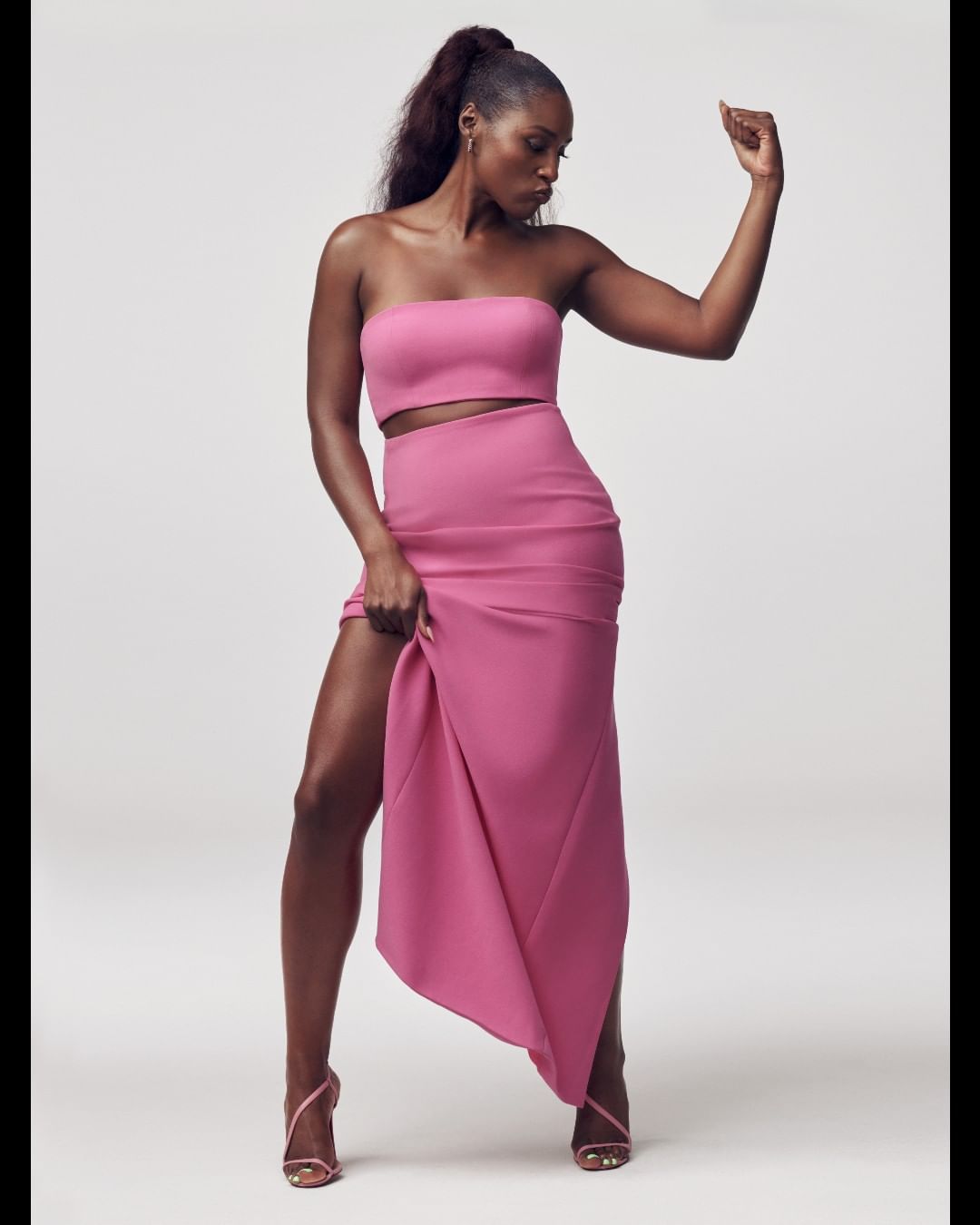 A confident woman in a pink two-piece dress and high heels poses against a clean background, displaying a raised arm and leg.