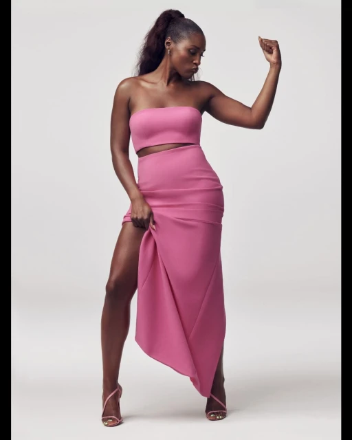 A confident woman in a pink two-piece dress and high heels poses against a clean background, displaying a raised arm and leg.