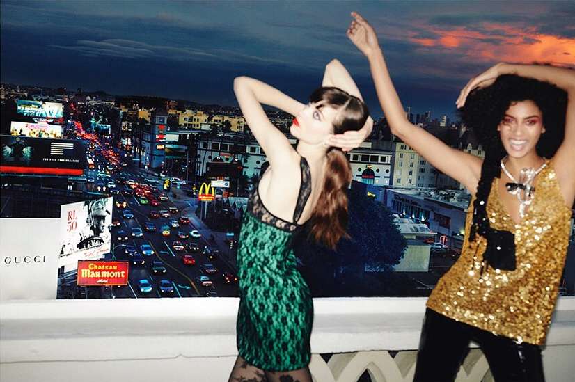 Two women in dresses pose in front of a billboard of Los Angeles city at night, featuring traffic, buildings, and sky.