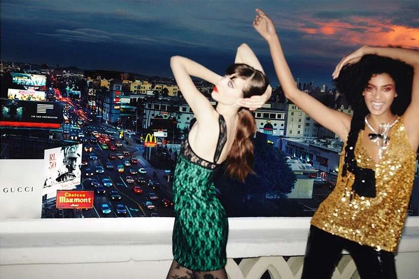 Two women in dresses pose in front of a billboard of Los Angeles city at night, featuring traffic, buildings, and sky.