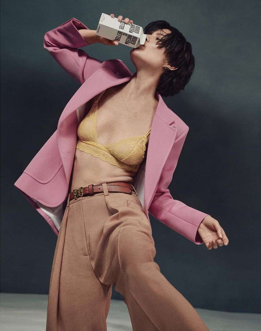 Brunette woman in bra and pink blazer drinking from a white beverage carton against a dark background in a fashion style pose.