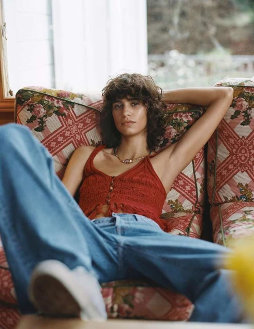 A woman with curly brown hair reclines on a floral couch, wearing a crocheted red top and blue jeans. The light is natural.