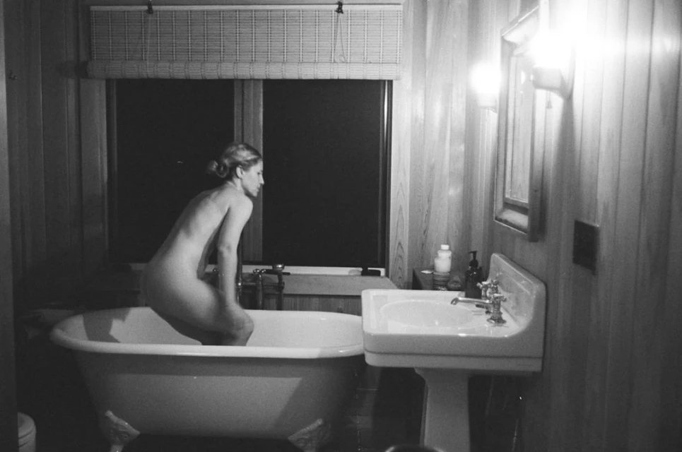Monochrome image of a woman perched on the edge of a clawfoot bathtub in a bathroom with wood paneling, a window, and a pedestal sink.