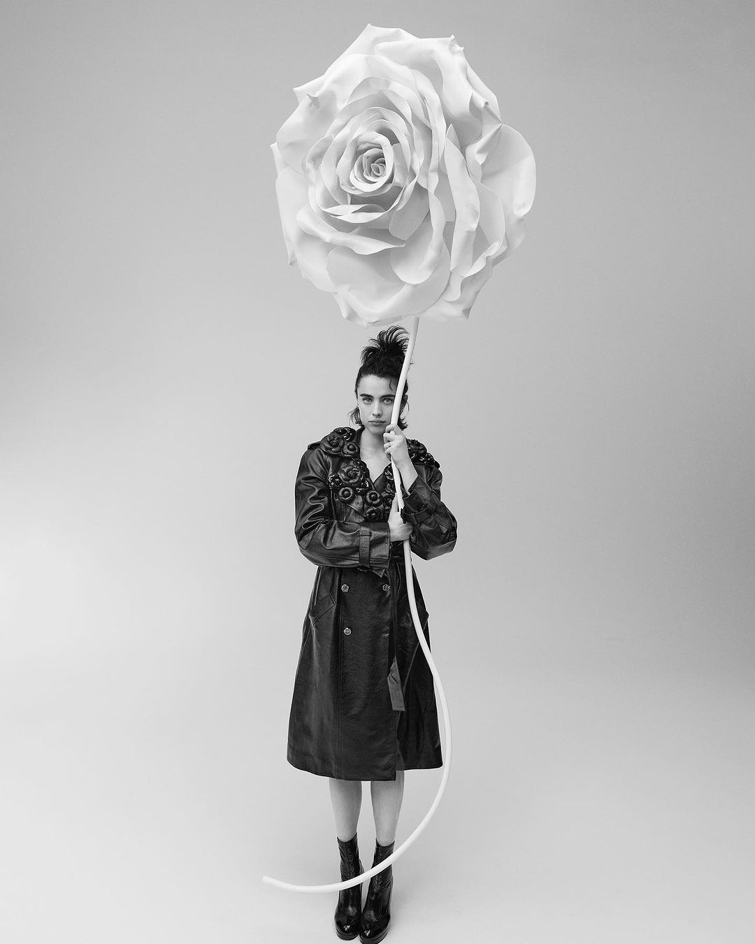 Monochrome portrait of a woman in a coat, holding an oversized rose, standing in a studio setting against a neutral background.