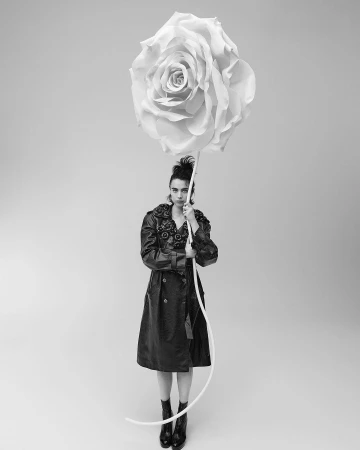 Monochrome portrait of a woman in a coat, holding an oversized rose, standing in a studio setting against a neutral background.