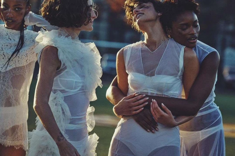 Four women in sheer white dresses pose outdoors, two of them embracing, against a blurred green and golden background, fashion photography.
