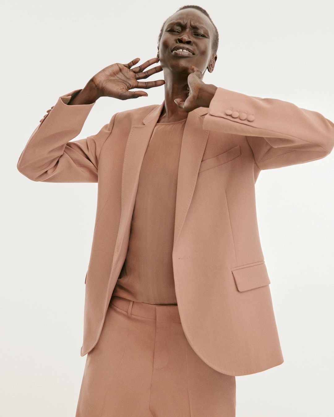 Model wearing a tan blazer and trousers with hands near neck against a seamless white background