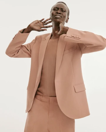 Model wearing a tan blazer and trousers with hands near neck against a seamless white background