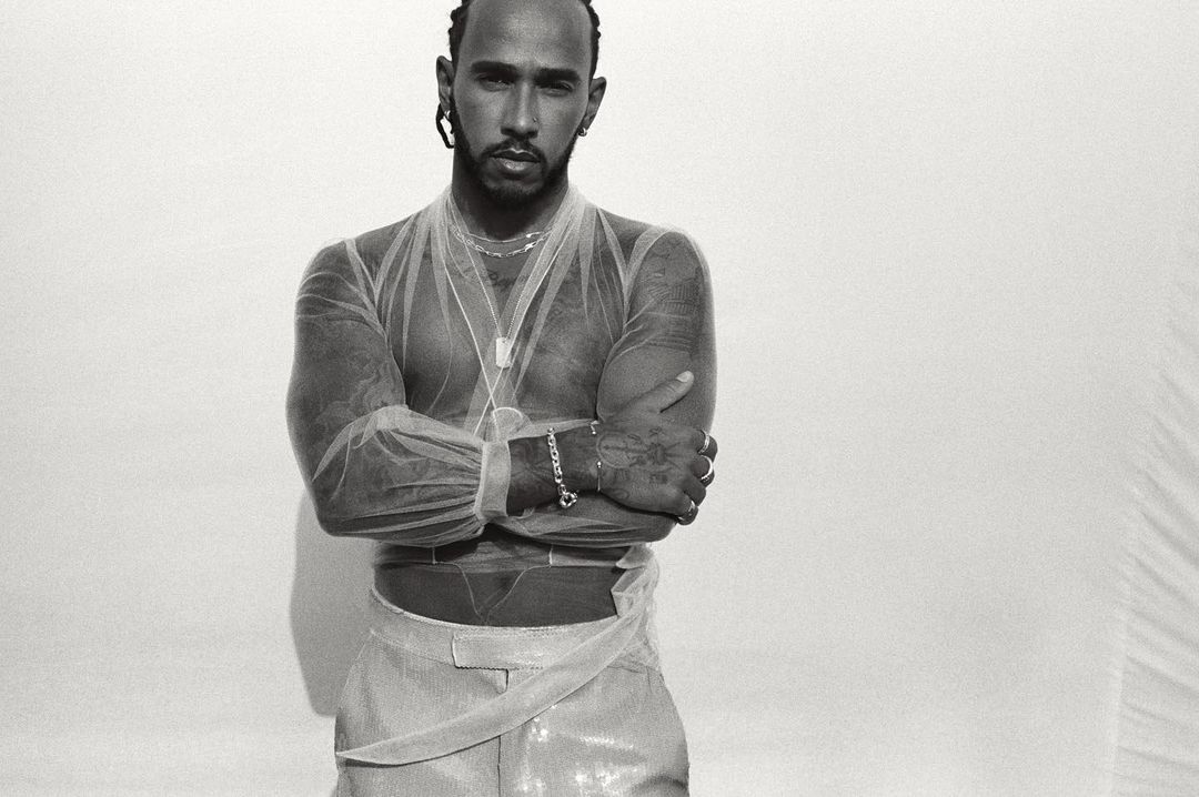 Portrait of Lewis Hamilton with arms crossed, wearing sheer top and shiny pants against a stark white backdrop in a photo studio.