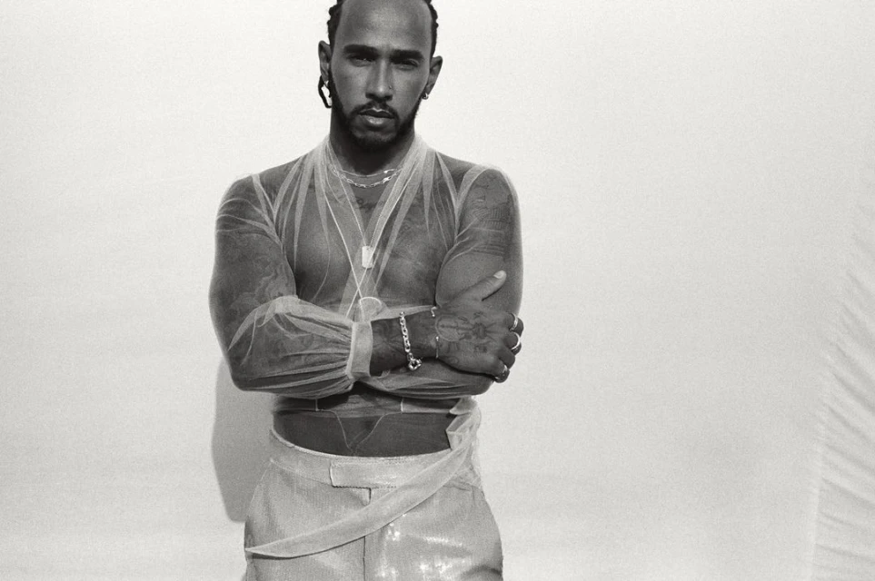 Portrait of Lewis Hamilton with arms crossed, wearing sheer top and shiny pants against a stark white backdrop in a photo studio.