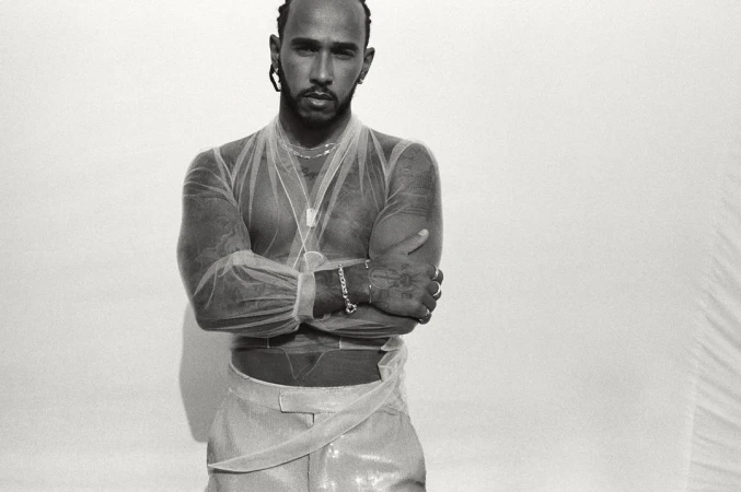 Portrait of Lewis Hamilton with arms crossed, wearing sheer top and shiny pants against a stark white backdrop in a photo studio.
