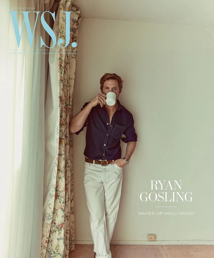 Ryan Gosling drinking from a white mug, wearing a dark blue shirt and white trousers against a plain wall.