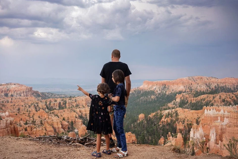 Father and two daughters stand at the edge of Bryce Canyon, looking out over the orange rock formations under a cloudy sky.