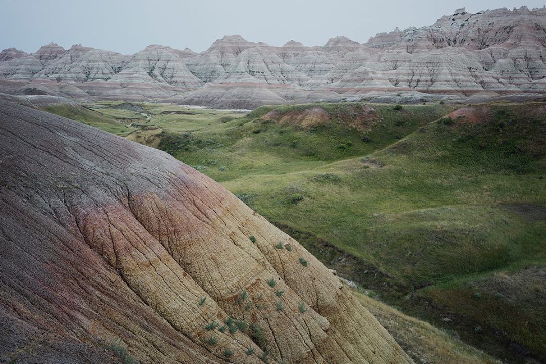 Scenic view of layered rock formations and grassy hills in Badlands National Park, South Dakota, under a cloudy sky, showcasing natural beauty.