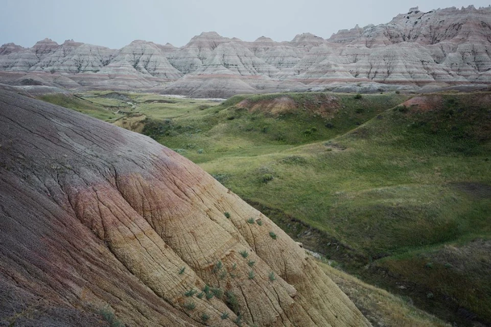 Scenic view of layered rock formations and grassy hills in Badlands National Park, South Dakota, under a cloudy sky, showcasing natural beauty.