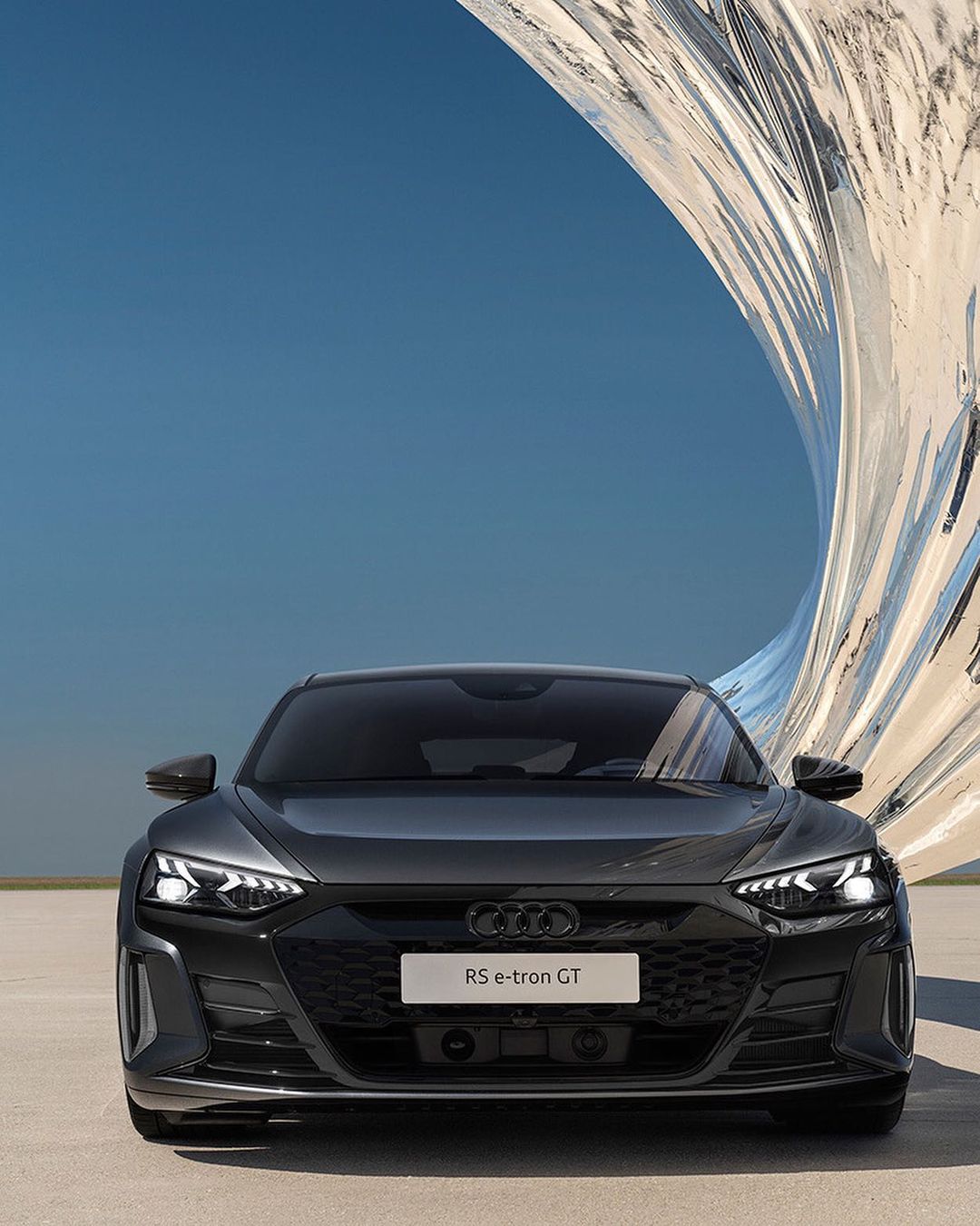 Front view of a dark gray Audi RS e-tron GT electric car parked on a concrete surface with a modern sculpture in the background.