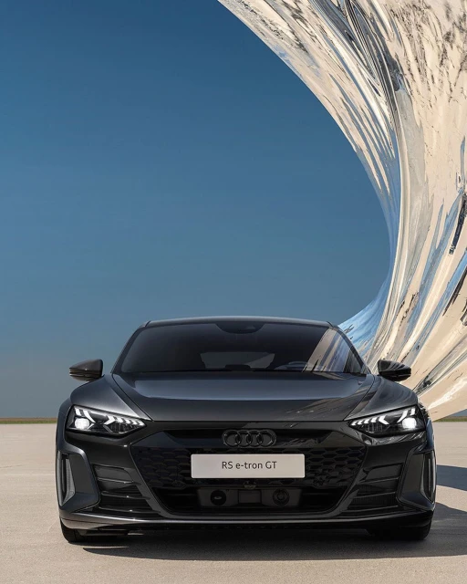 Front view of a dark gray Audi RS e-tron GT electric car parked on a concrete surface with a modern sculpture in the background.