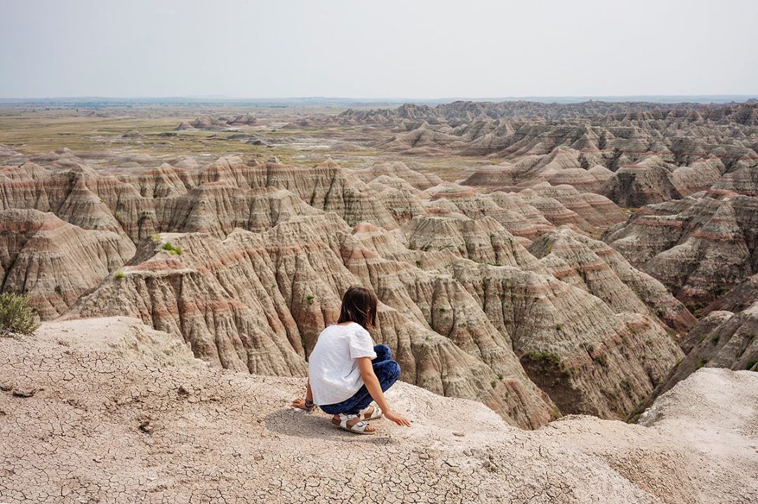 Young woman crouches on a plateau overlooking the vast, striated landscape of Badlands National Park on a cloudy day.