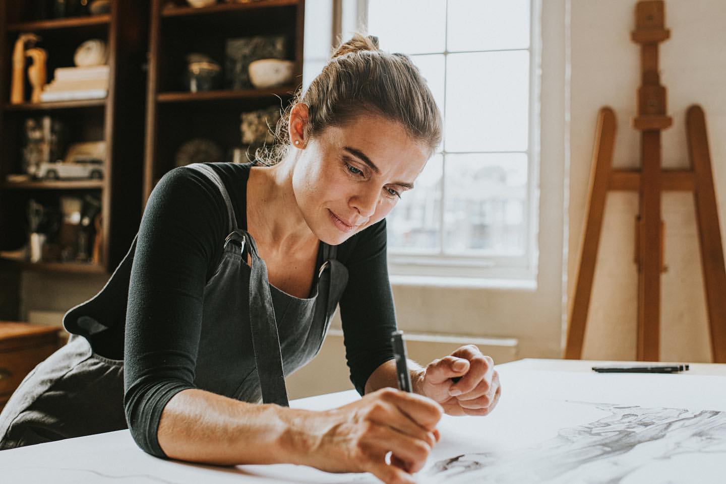 Adult female artist in an apron leaning over a drawing at a desk in a brightly lit studio with a wooden easel and shelves.