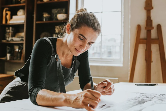 Adult female artist in an apron leaning over a drawing at a desk in a brightly lit studio with a wooden easel and shelves.