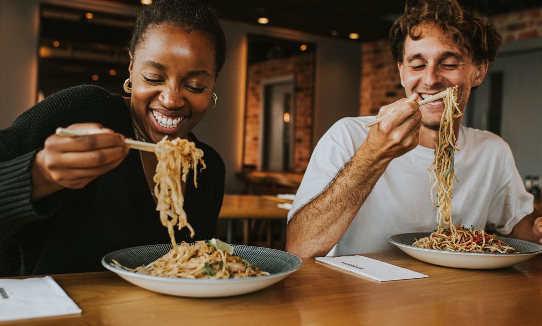 A smiling woman and man enjoy noodles with chopsticks at a table, capturing a joyful dining experience in a warm, inviting setting.