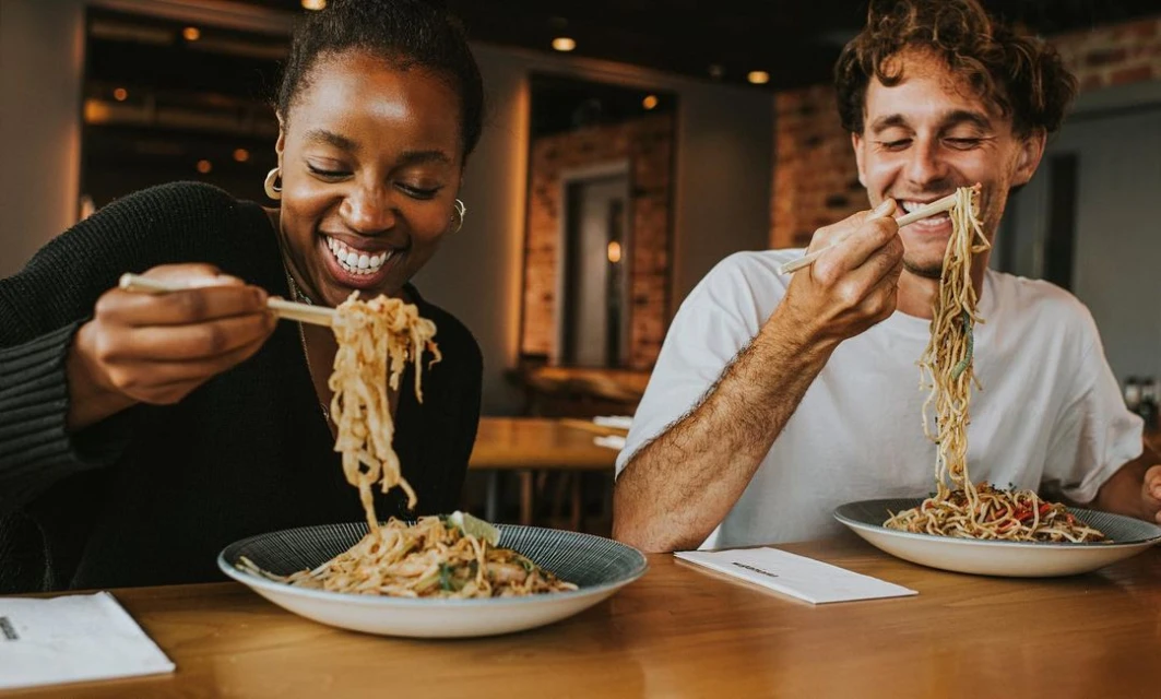 A smiling woman and man enjoy noodles with chopsticks at a table, capturing a joyful dining experience in a warm, inviting setting.