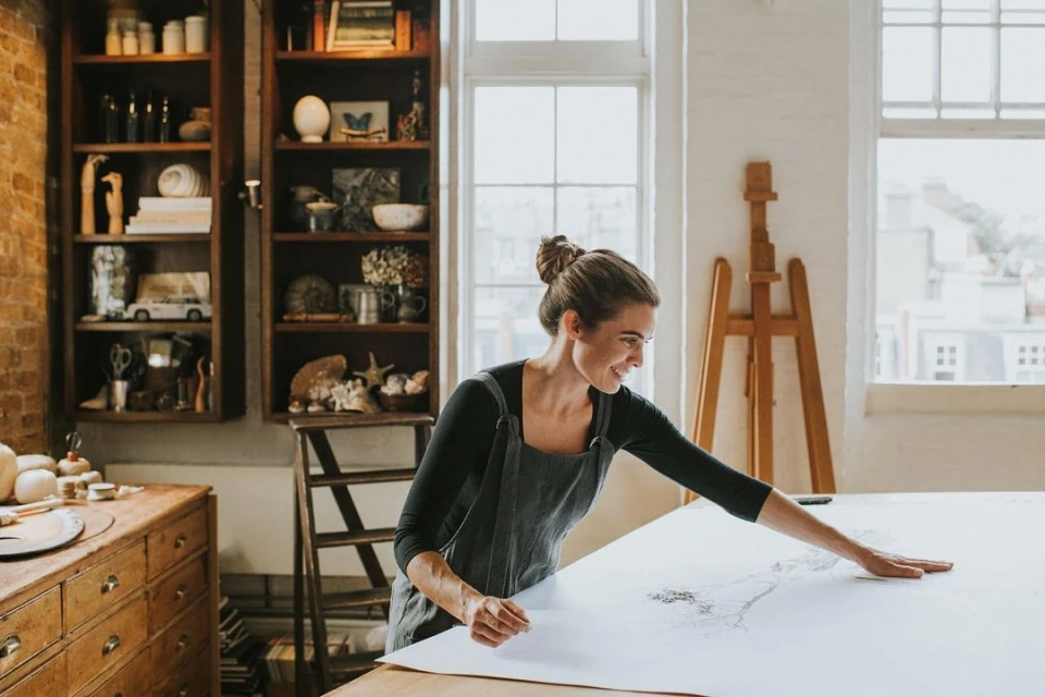 A woman in a grey apron smiles as she works on a large drawing on a wooden table in a bright artist studio with natural light.
