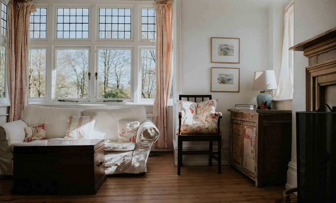 Sunlit living room featuring a white sofa, wooden table, floral cushions, a window overlooking trees, and vintage decor details.