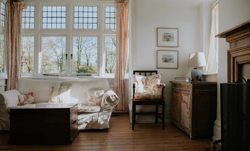 Sunlit living room featuring a white sofa, wooden table, floral cushions, a window overlooking trees, and vintage decor details.