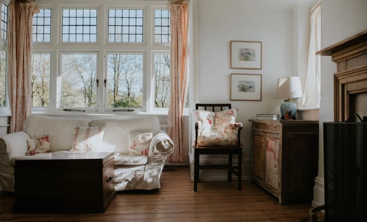 Sunlit living room featuring a white sofa, wooden table, floral cushions, a window overlooking trees, and vintage decor details.