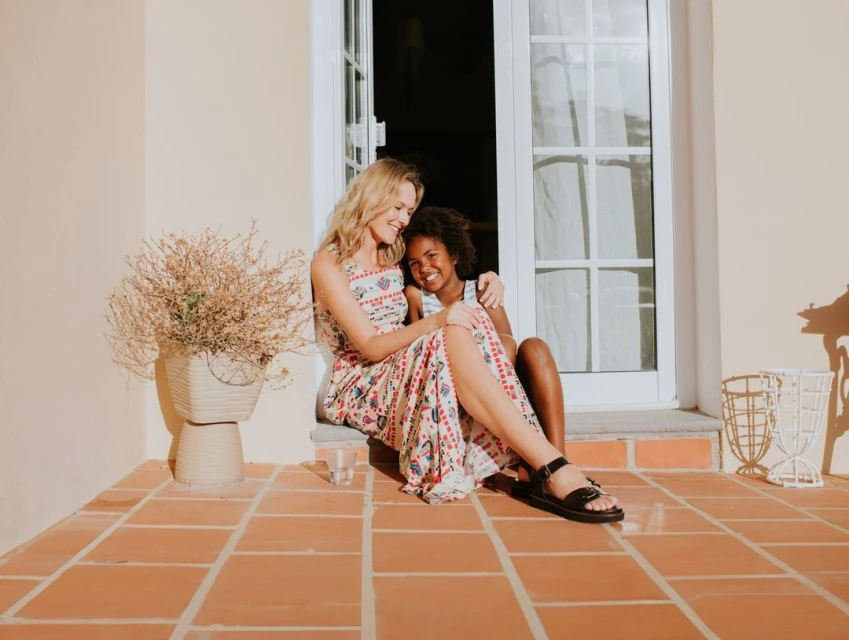 Blonde woman and an African-American child sitting on steps of a building, embracing in natural light with a plant and window behind them.