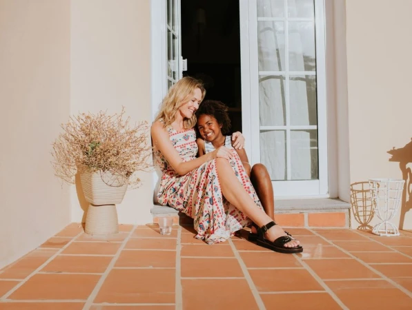Blonde woman and an African-American child sitting on steps of a building, embracing in natural light with a plant and window behind them.