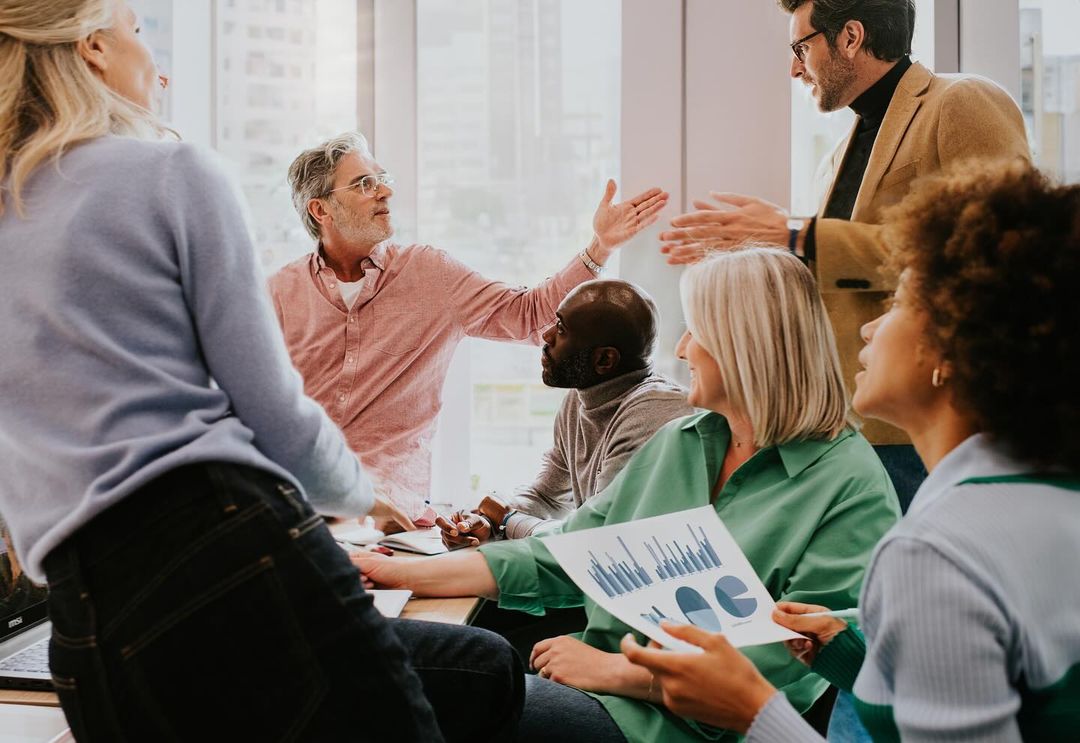 Group of diverse business professionals collaborate in a modern office setting, reviewing charts and graphs, and engaging in discussion.