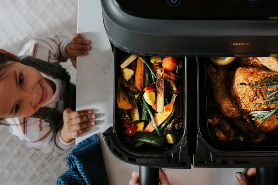 Child smiles next to an air fryer filled with roasted vegetables and cooked chicken, illuminated by indoor kitchen lighting.