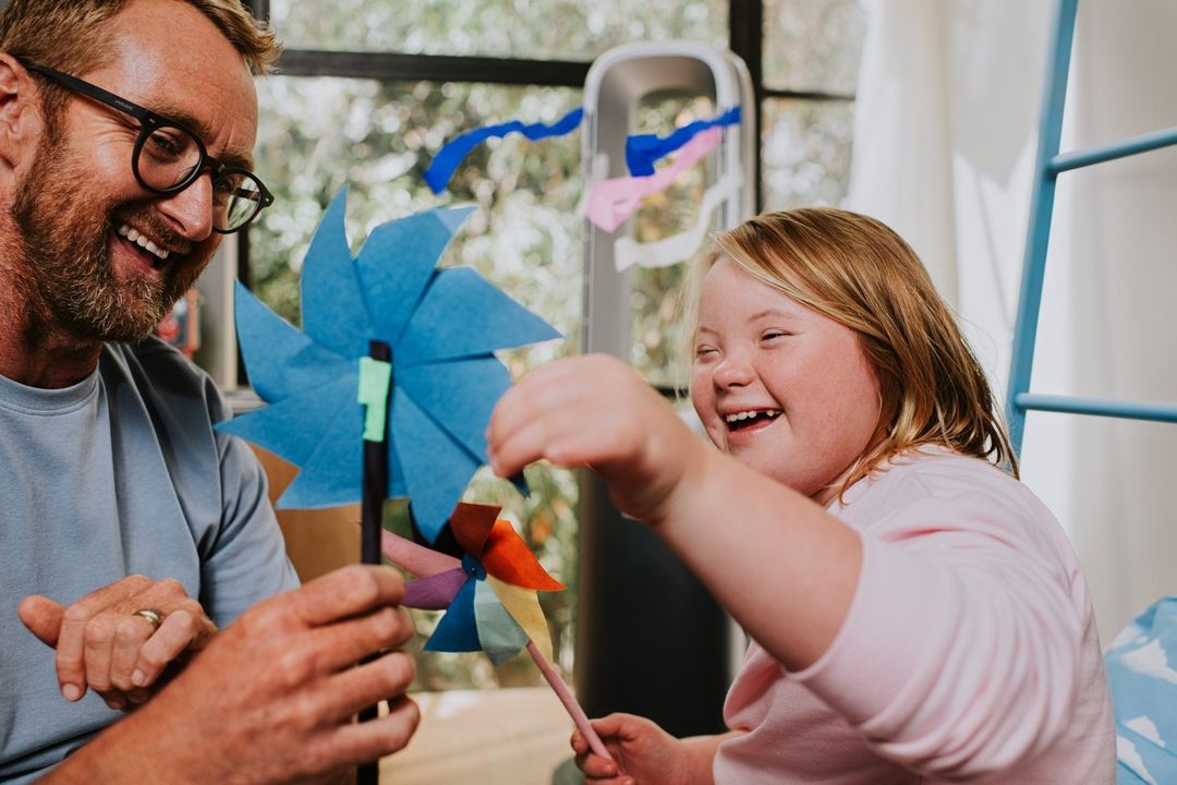 Smiling father and daughter create a colorful paper pinwheel indoors, bathed in soft natural light, exuding a heartwarming, cheerful mood.