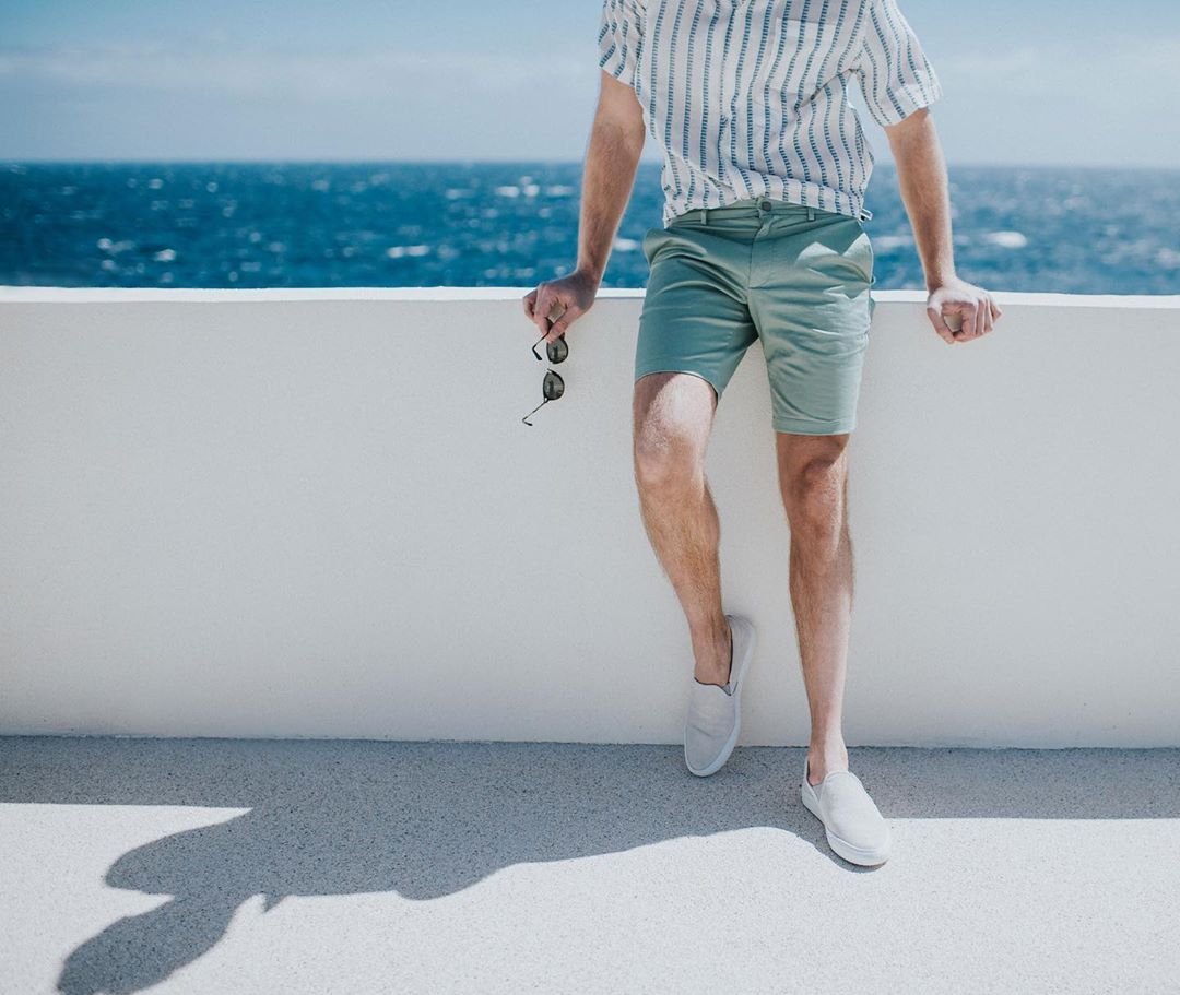 An adult male in shorts and a striped shirt leans against a white wall, holding sunglasses, with a sea backdrop under a clear sky.