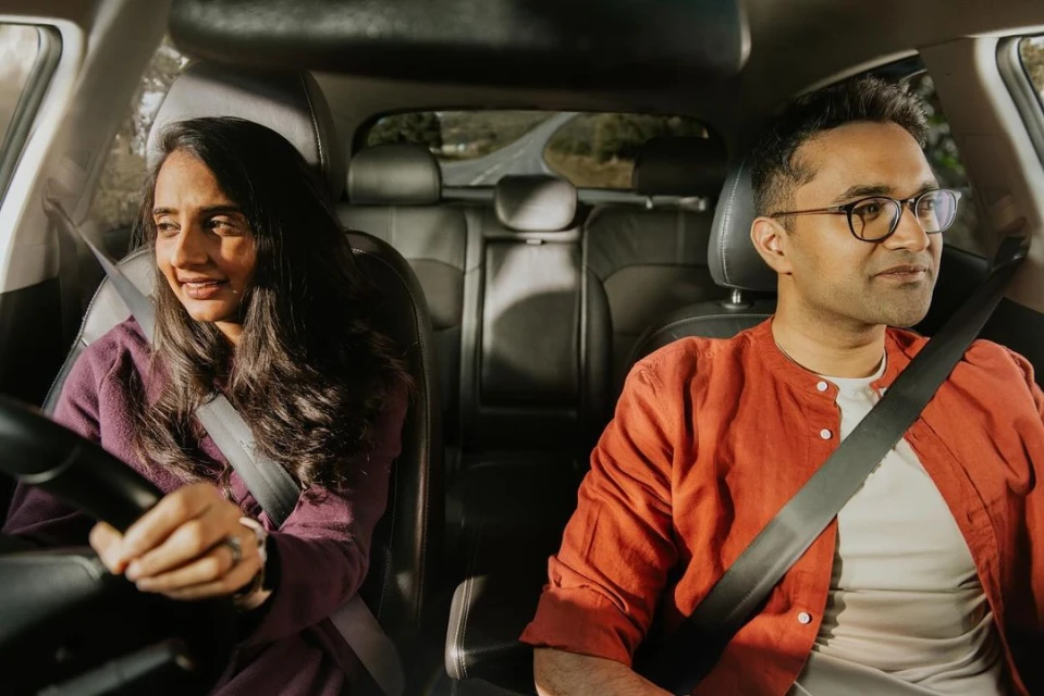 Woman driving a car with a male passenger. The couple is on a road trip with trees visible through the windows of the car.