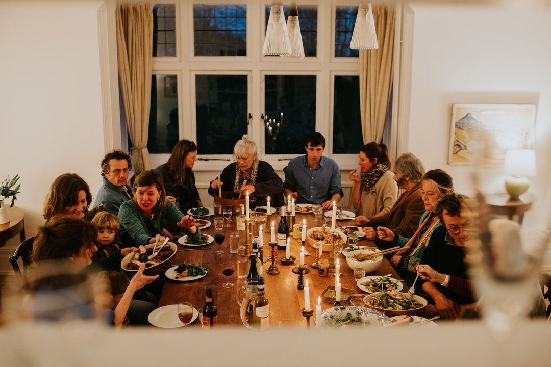 Candid shot of a diverse group of people around a long wooden table with candles, plates of food, and wine bottles in a home interior.
