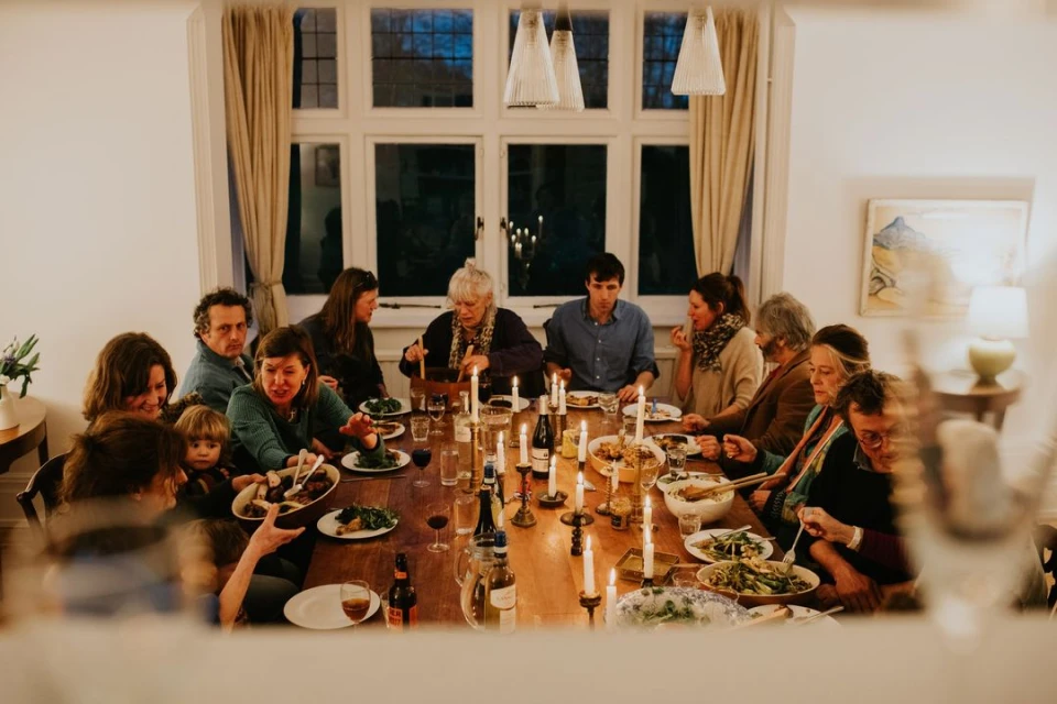 Candid shot of a diverse group of people around a long wooden table with candles, plates of food, and wine bottles in a home interior.